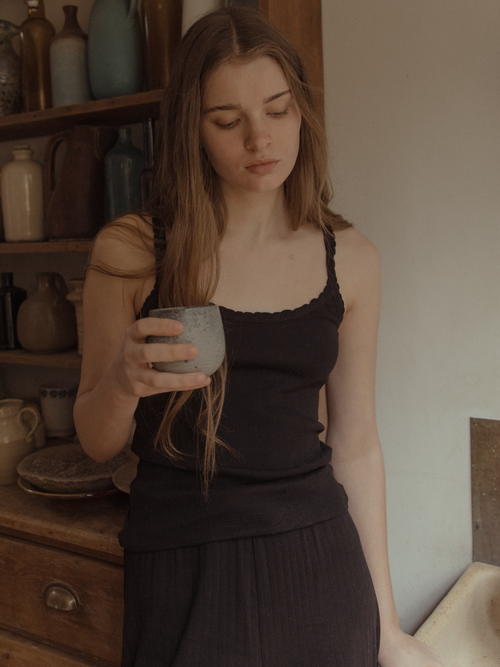Model wearing the Calla Tank in Noir by Gillian Stevens, holding a ceramic cup in a rustic kitchen setting