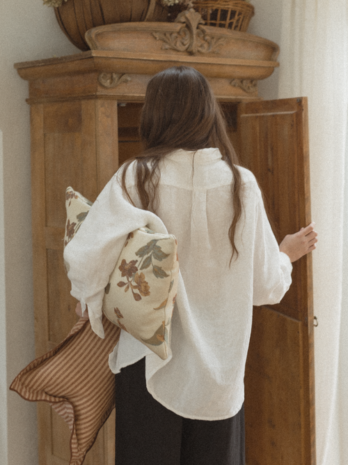 Woman holding pillows in front of a wooden wardrobe wearing a Britta Shirt Linen Salt by Gillian Stevens.
