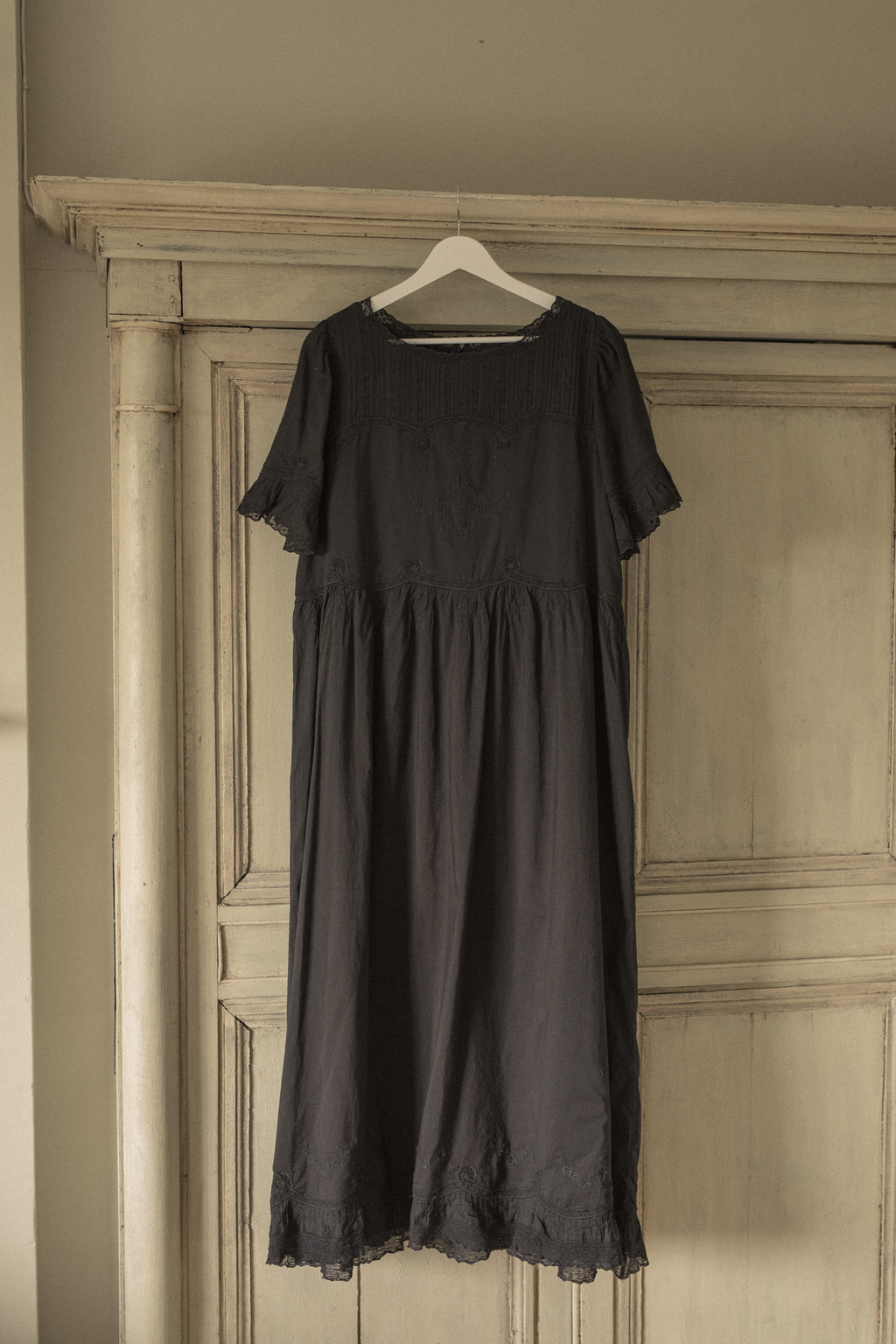 Black dress, the Astrid dress in noir  by Gillian Stevens, hanging on a hanger against a door frame
