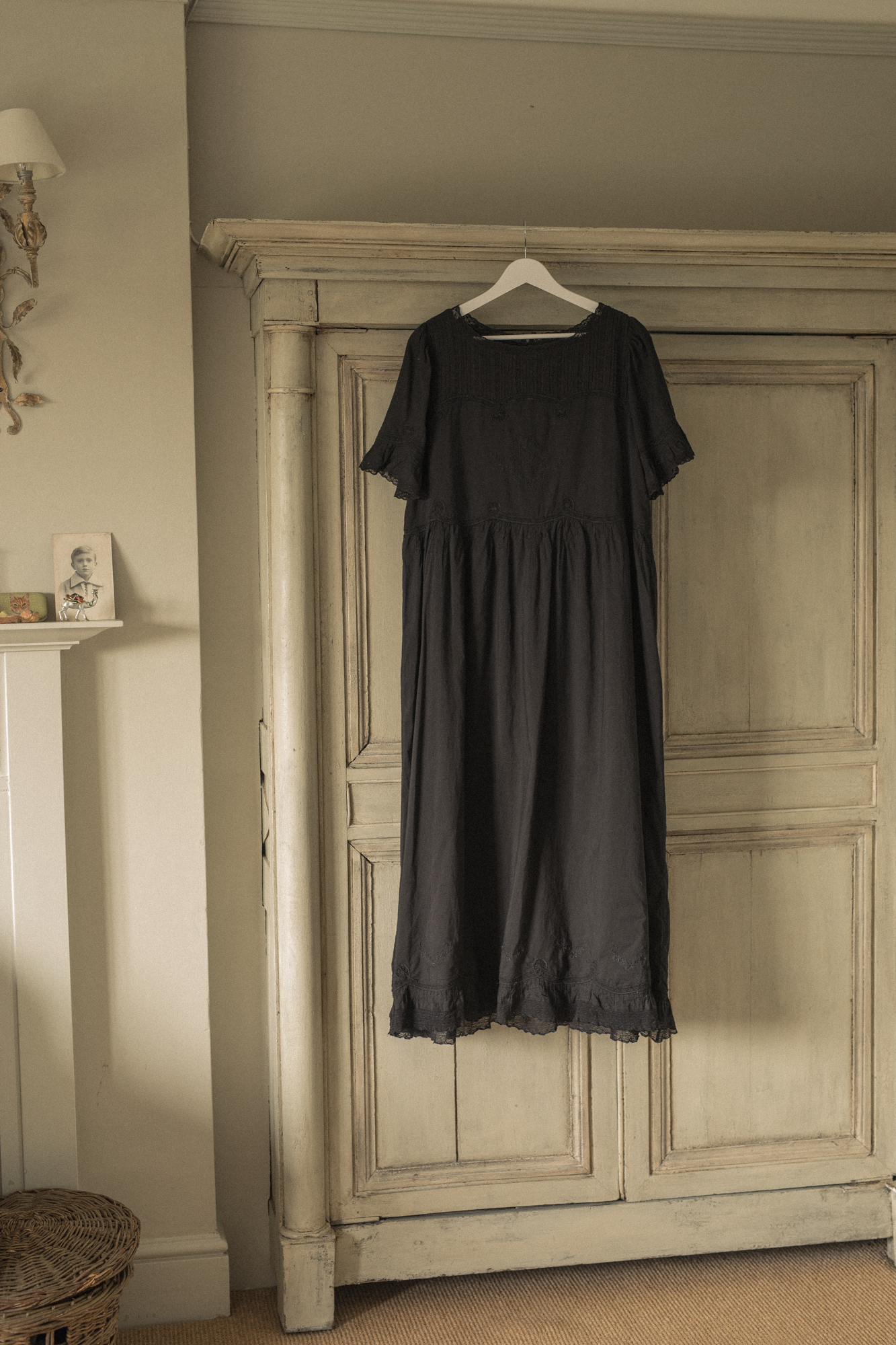 Black dress, the Astrid dress in noir by Gillian Stevens, hanging on a wooden wardrobe in a room with neutral walls.