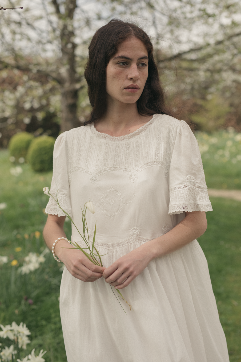 Woman in a white dress holding grass in a garden wearing the astrid dress in salt by Gillian Stevens