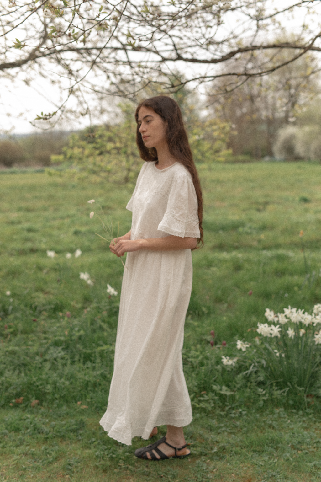 Woman in a white dress standing in a grassy field with trees and flowers wearing the astrid dress in salt by Gillian Stevens