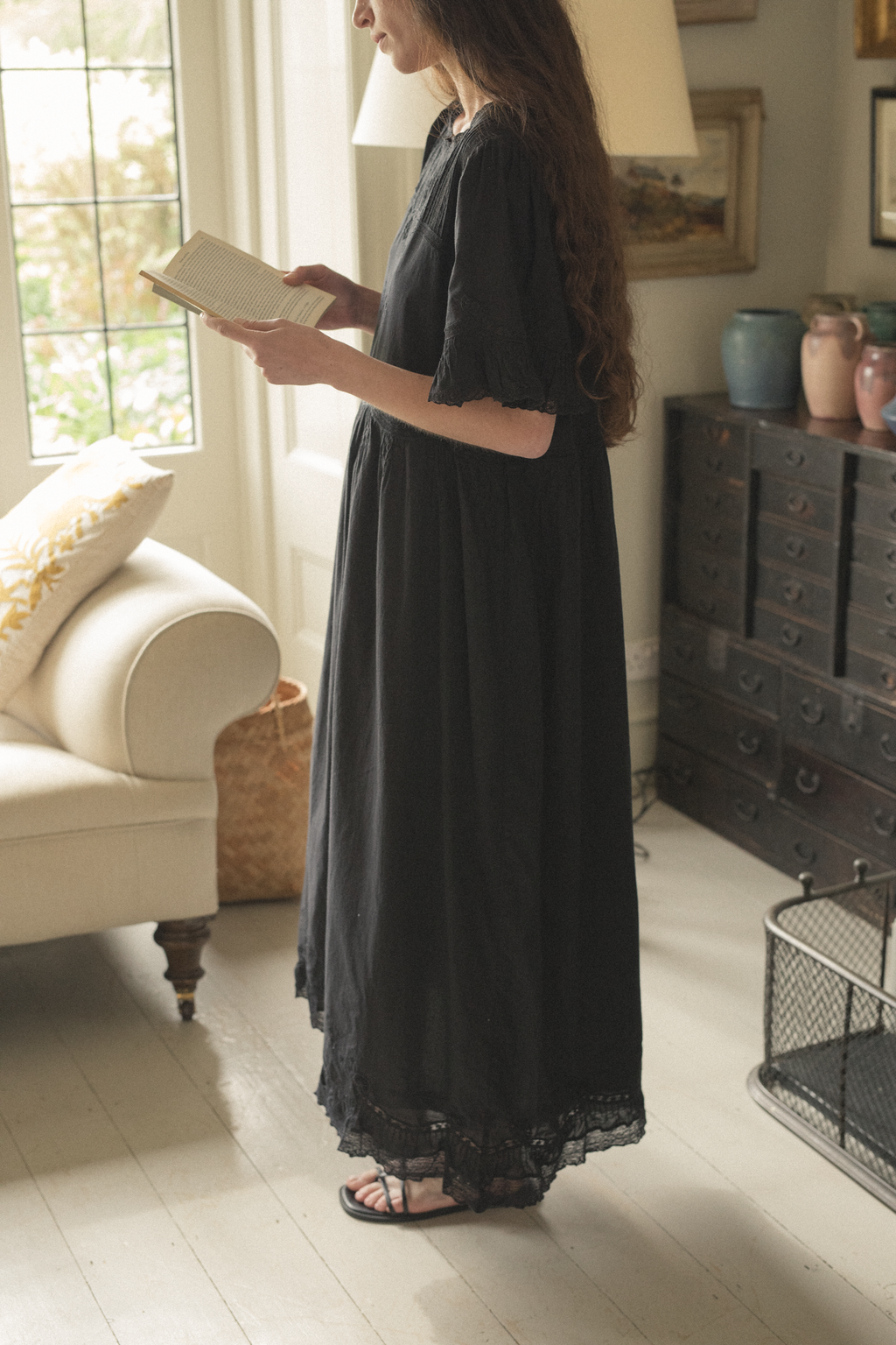 Woman in a black dress, the Astrid dress in noir by Gillian Stevens, reading a book in a cozy living room.