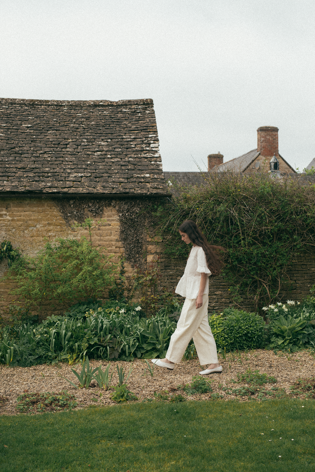 A woman walking in a garden with a rustic building in the background, wearing the Astrid blouse in salt by Gillian Stevens