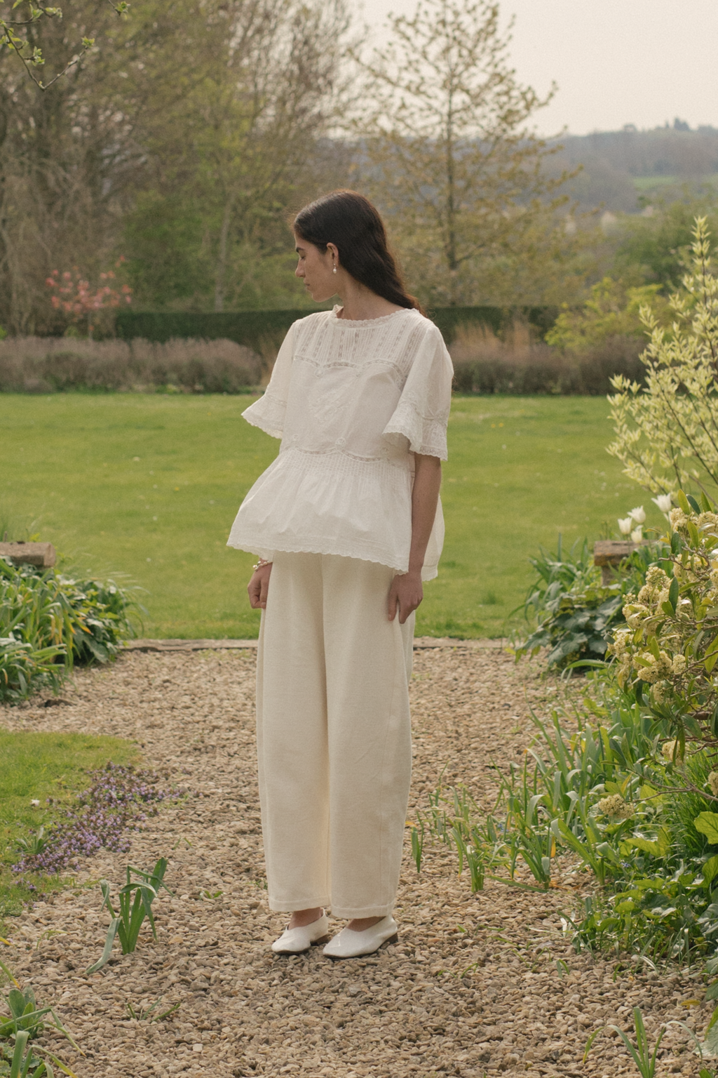 Woman in a white blouse with lace and  embroidery, the Astrid blouse in salt by Gillian Stevens, standing in a garden