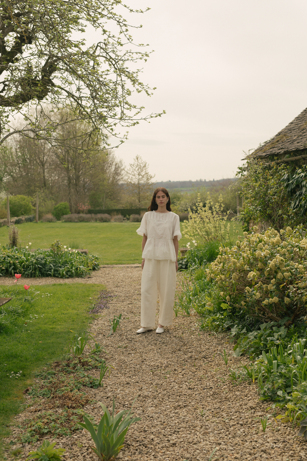 Woman in a white top, the Astrid blouse in salt by Gillian Stevens, and pant standing on a gravel path in a garden.