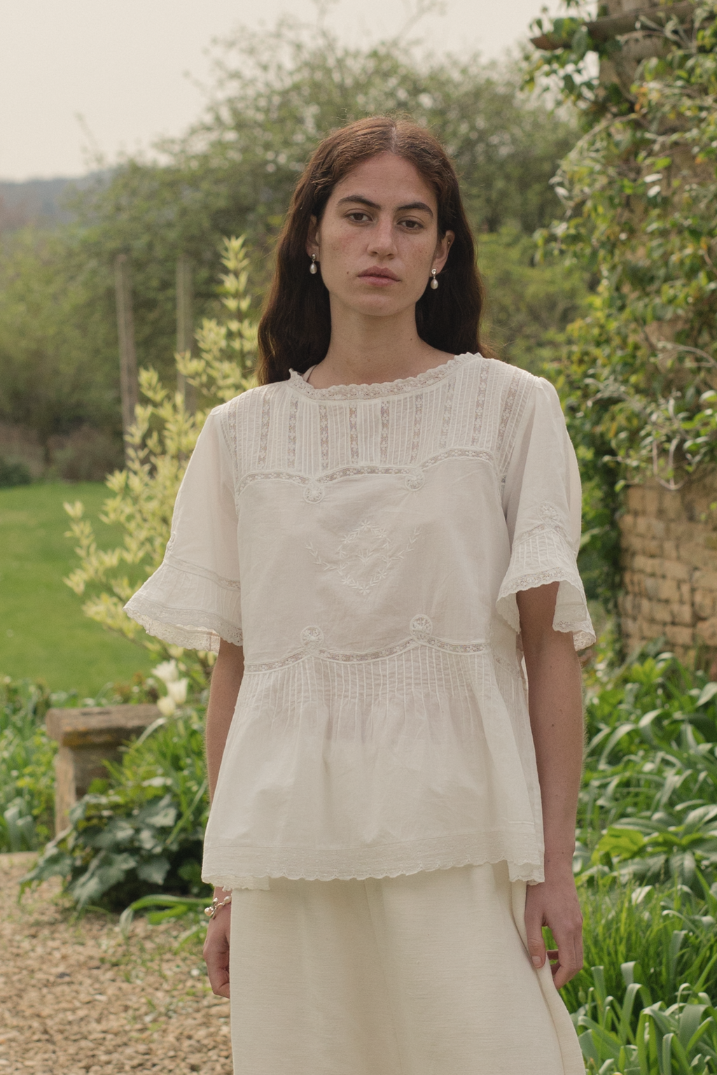 Woman wearing a white embroidered cotton blouse, the Astrid blouse in salt by Gillian Stevens, and pants in a garden setting