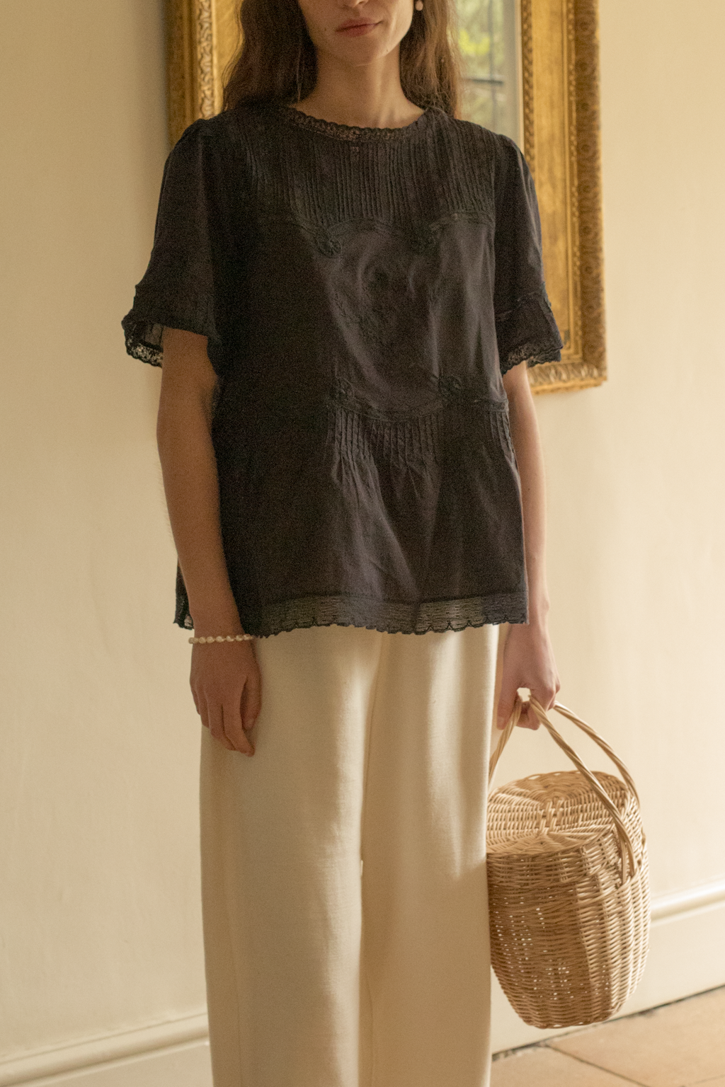 Woman wearing a black lace blouse, the Astrid top in noir by Gillian Stevens, and white pants, holding a woven bag indoors.