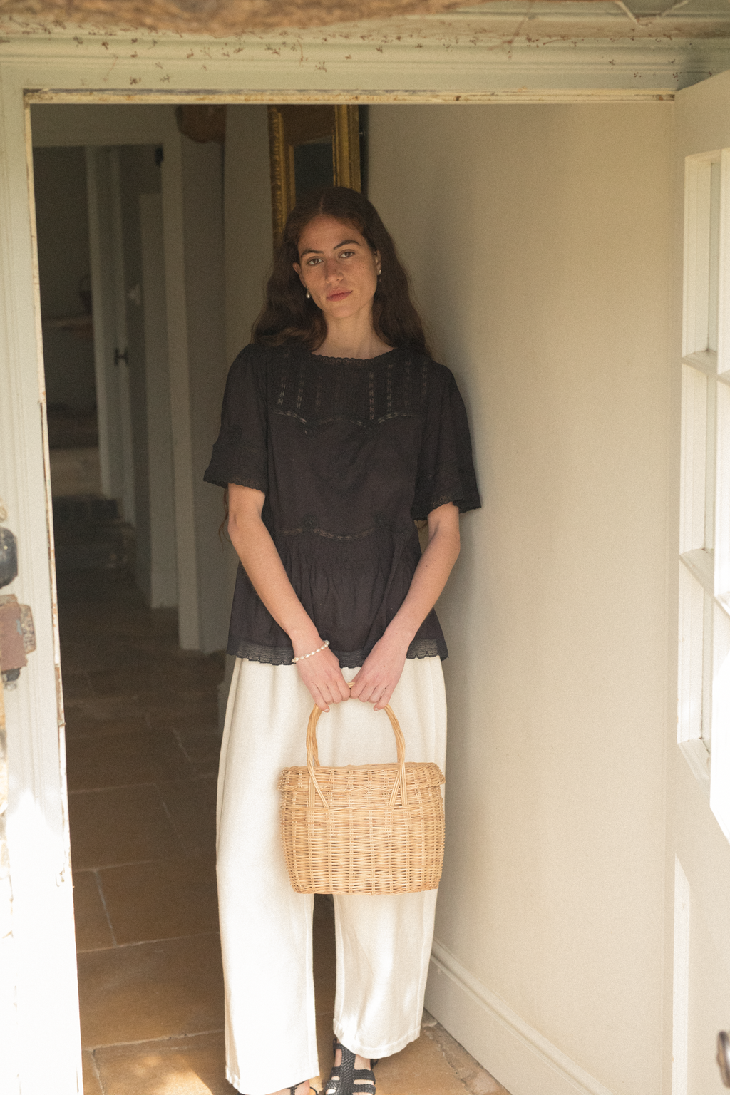 Woman holding a woven basket in a room with a wooden floor and white walls wearing the Astrid Blouse in noir by Gillian Stevens