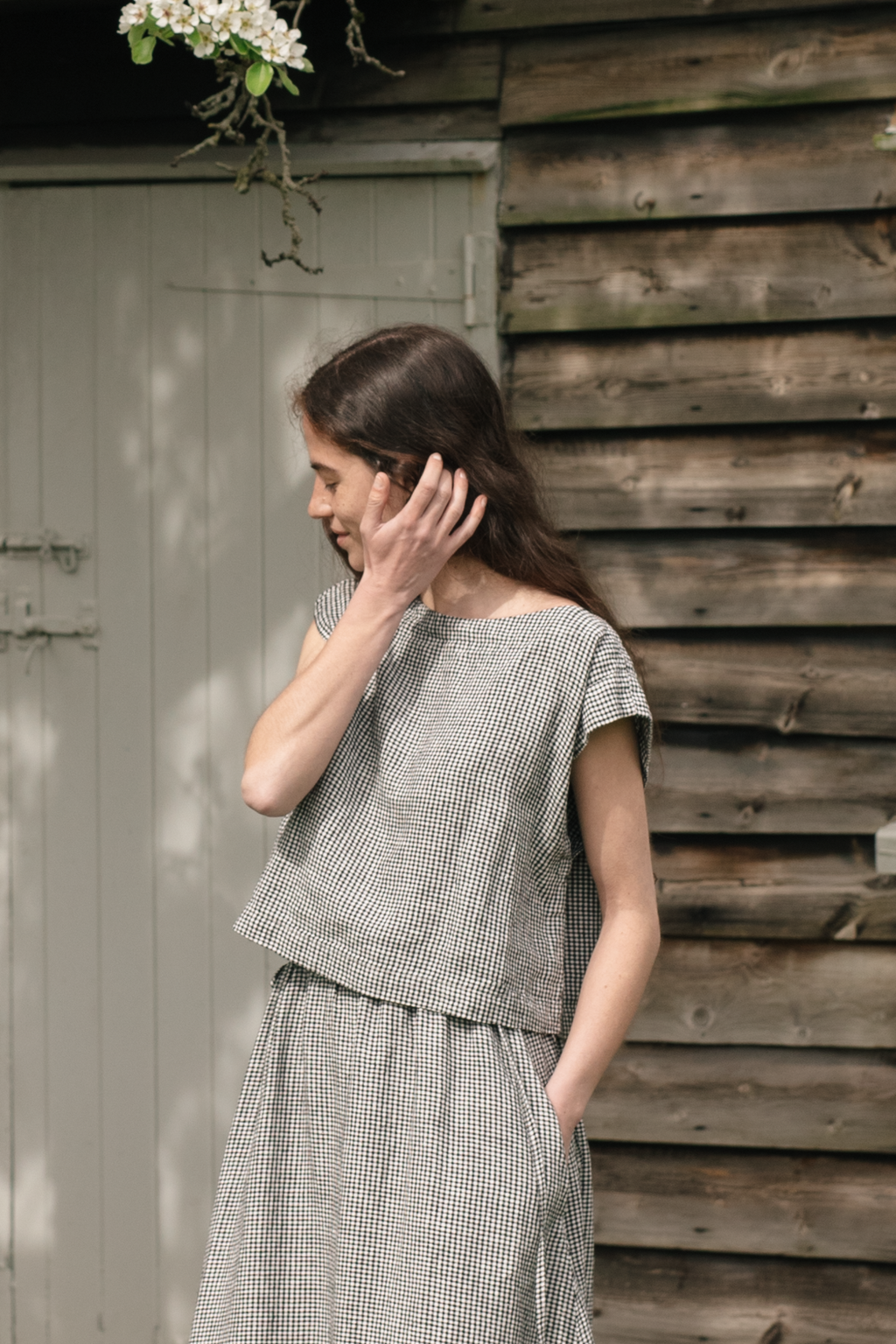 Woman wearing a checkered top, the Valley top in Gingham Noir by Gillian Stevens, and skirt standing against a wooden wall.