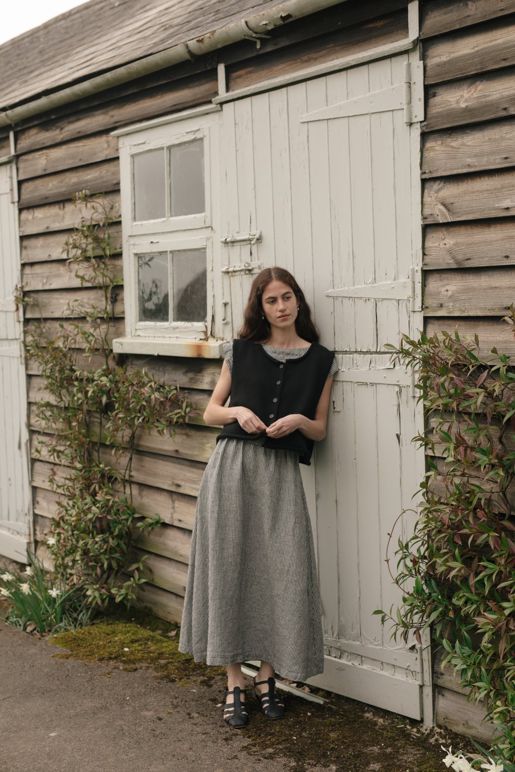 Model wearing the Lennon Vest in Noir by Gillian Stevens, full-length outdoor shot against a rustic wooden shed