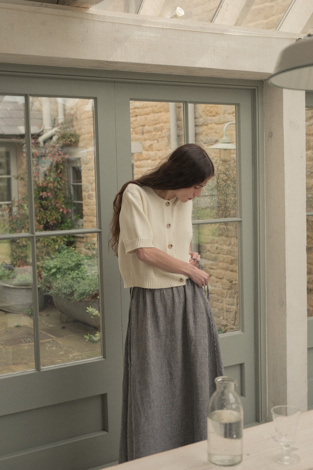 Woman standing in a room with large windows, looking outside, wearing the rosemary cardigan in alabaster by Gillian Stevens,