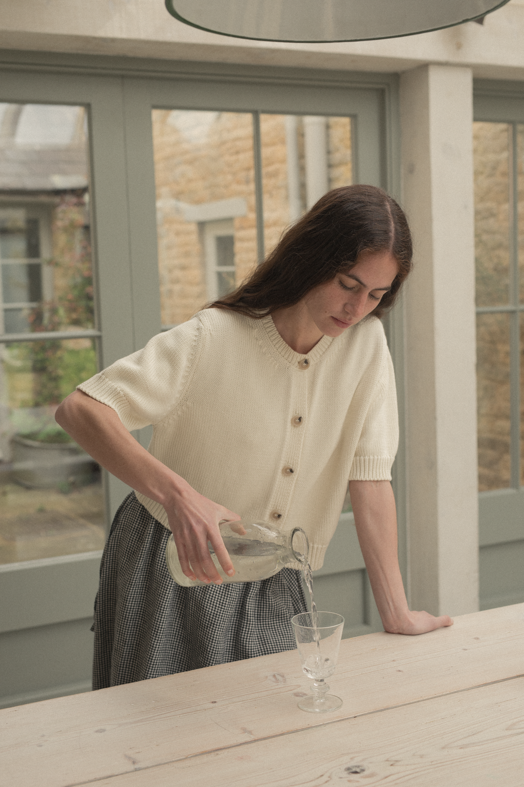 Model wearing the Rosemary Cardigan in Alabaster by Gillian Stevens, leaning over a table pouring water