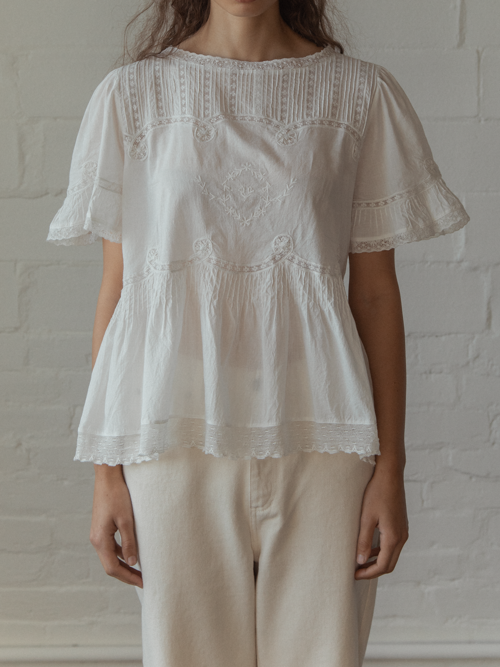 Woman wearing a white lace blouse, the Astrid blouse in salt by Gillian Stevens, against a white brick wall.