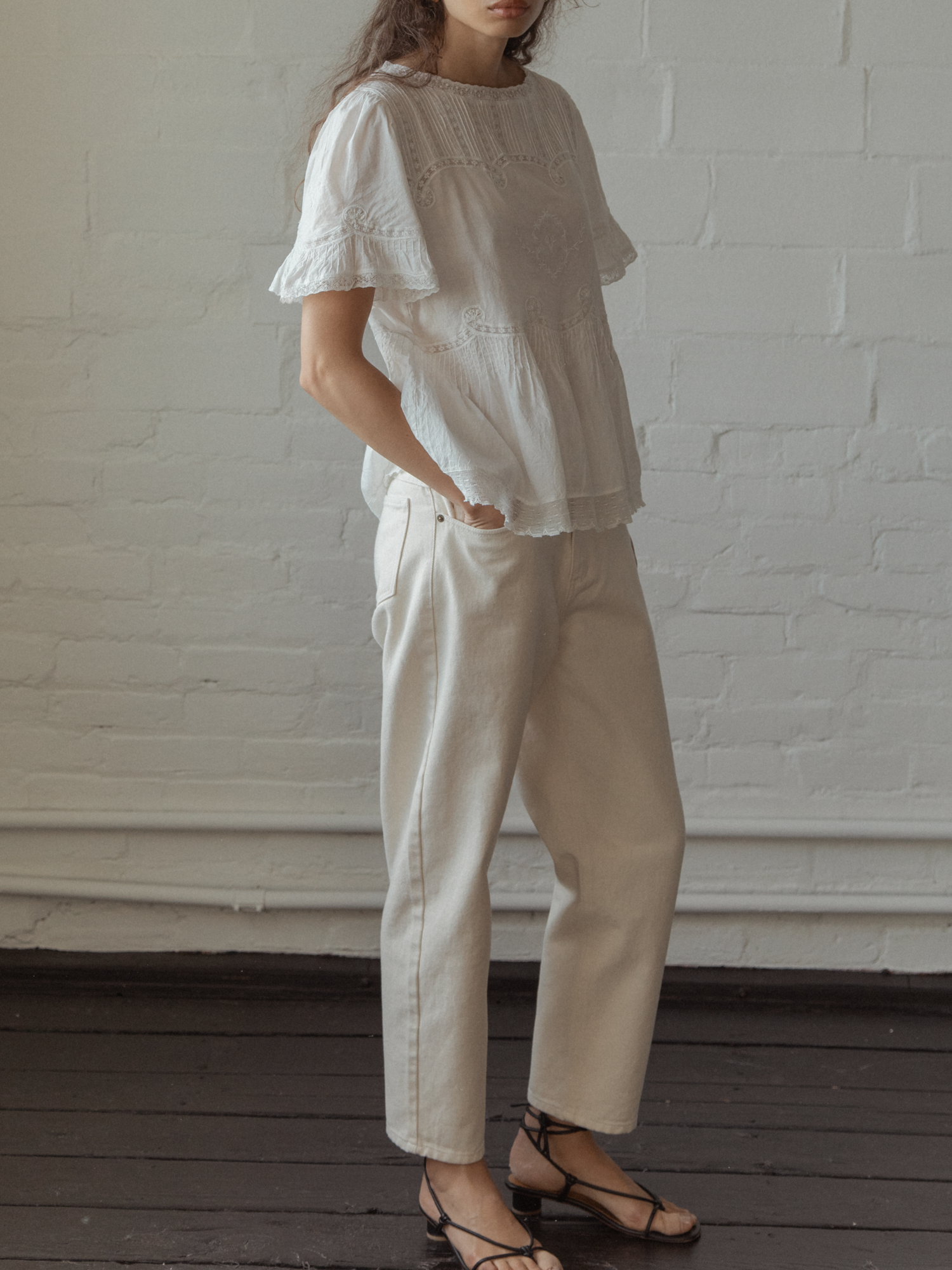 A Woman wearing a white top, the Astrid blouse in salt by Gillian Stevens,  and beige pants against a white brick wall.