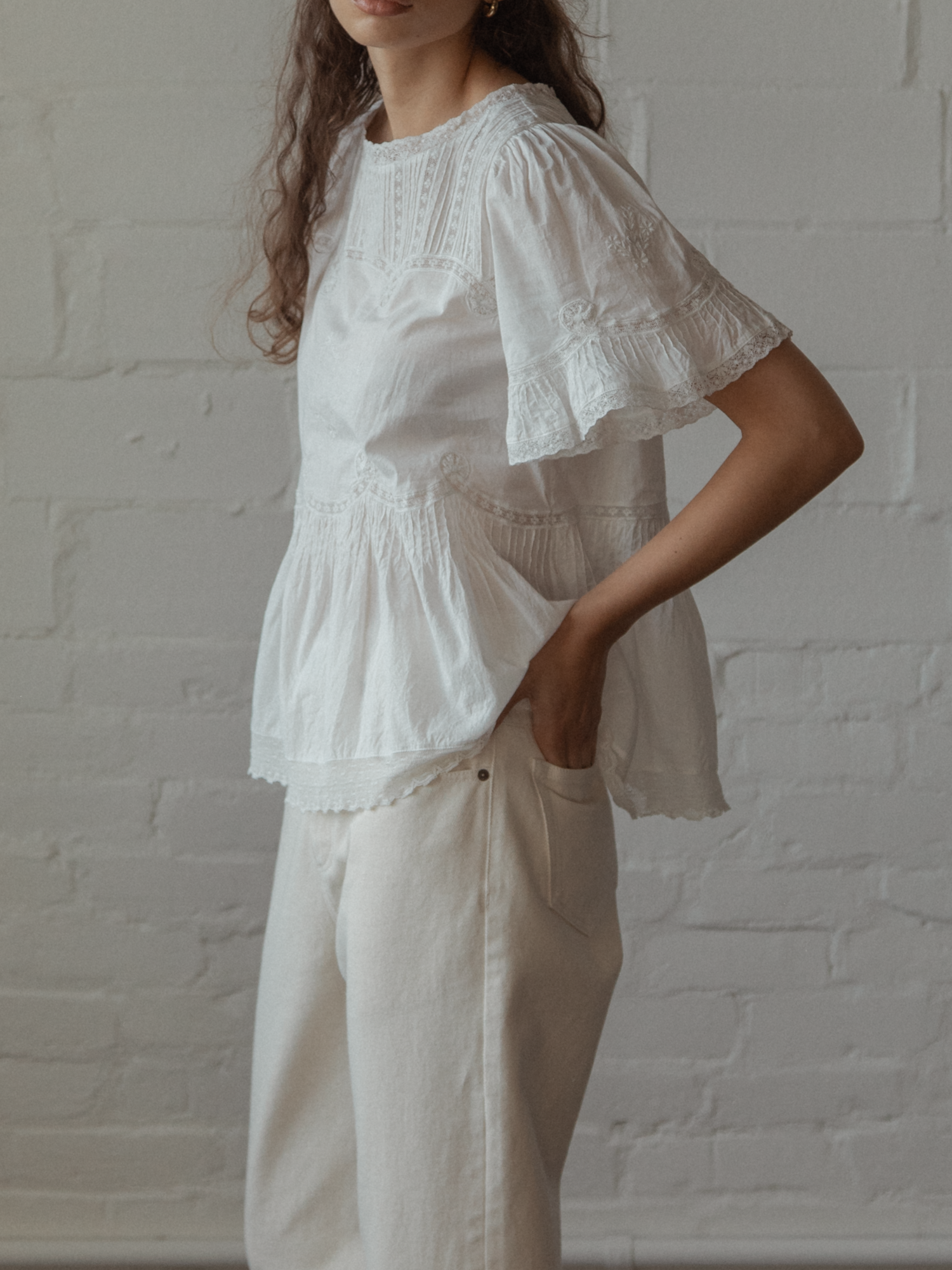 Woman wearing a white lace top, the Astrid blouse in salt by Gillian Stevens, and beige pants against a white brick wall.