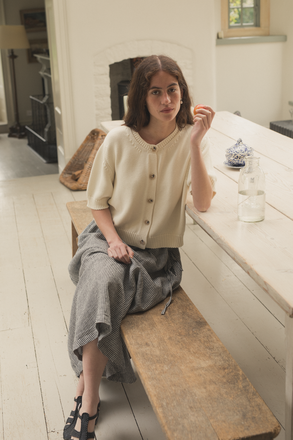 Woman sitting on a wooden bench in a kitchen wearing the rosemary cardigan in alabaster by Gillian Stevens,