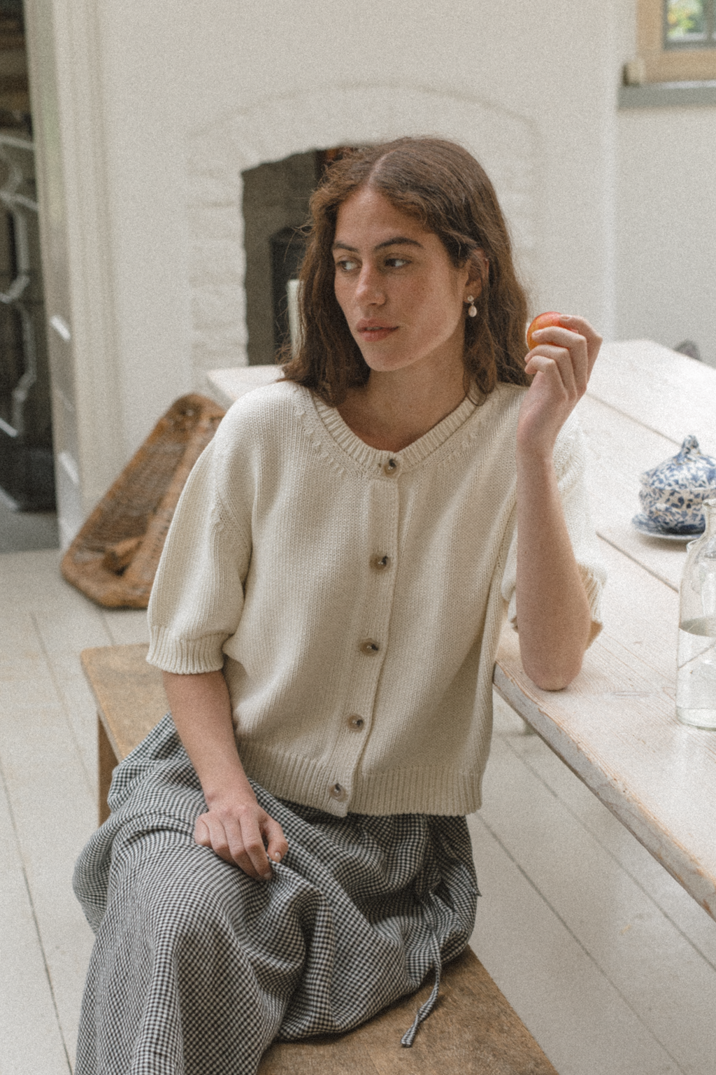 Model wearing the Rosemary Cardigan in Alabaster by Gillian Stevens, seated front view in a rustic interior