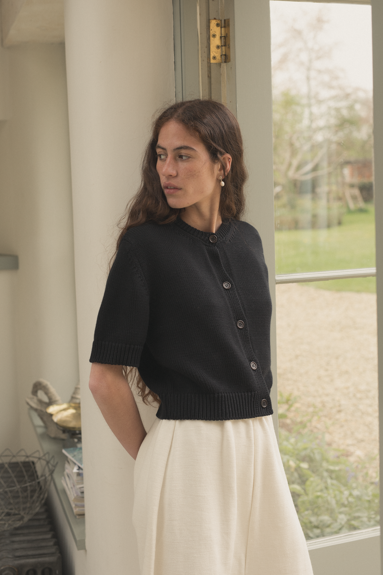 Woman wearing a black cardigan, the rosemary cardigan by  Gillian Stevens, standing indoors with a window in the background