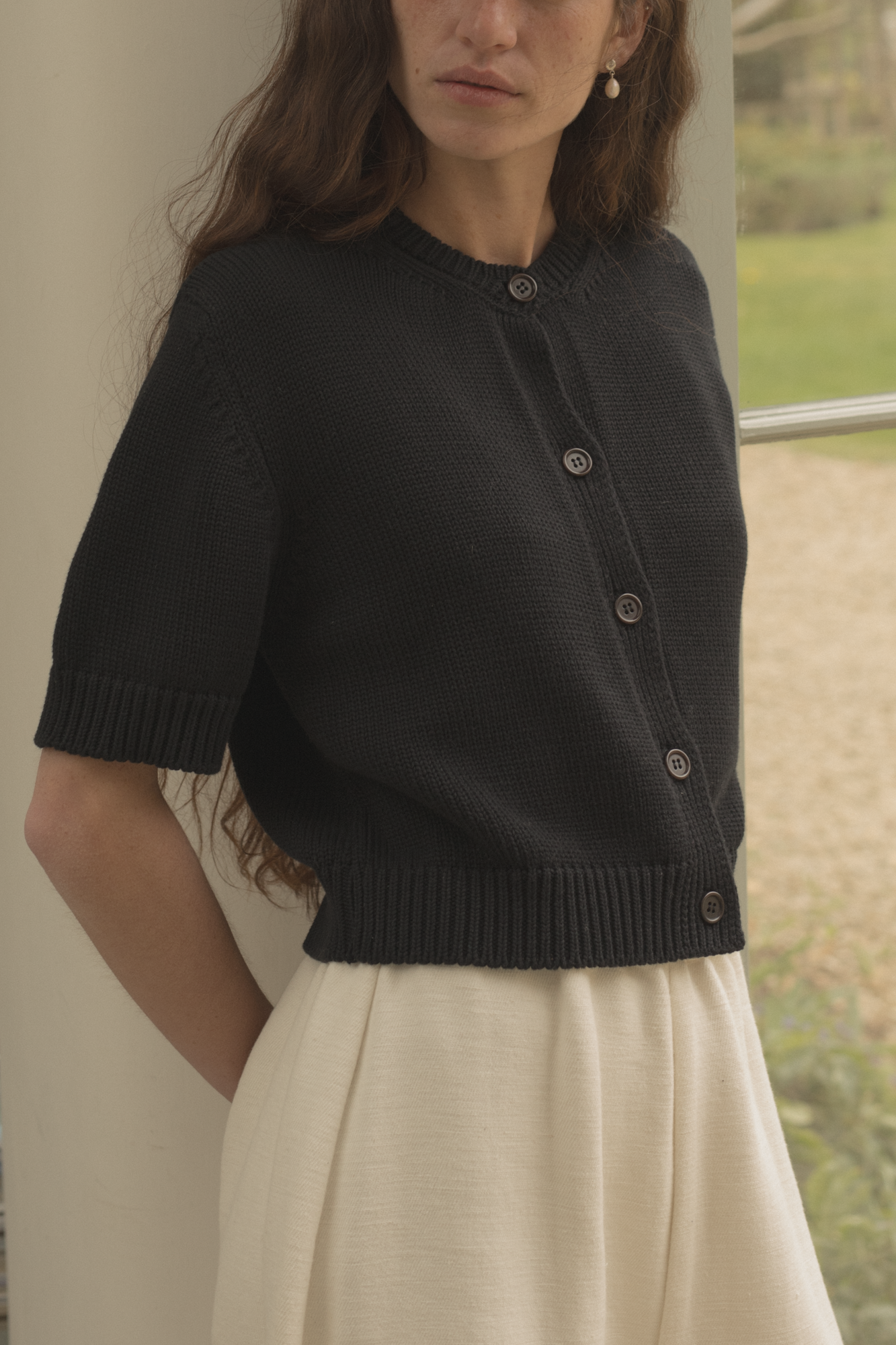 Woman wearing a black cardigan, the rosemary cardigan in Noir by Gillian Stevens, and white pants standing outdoors.