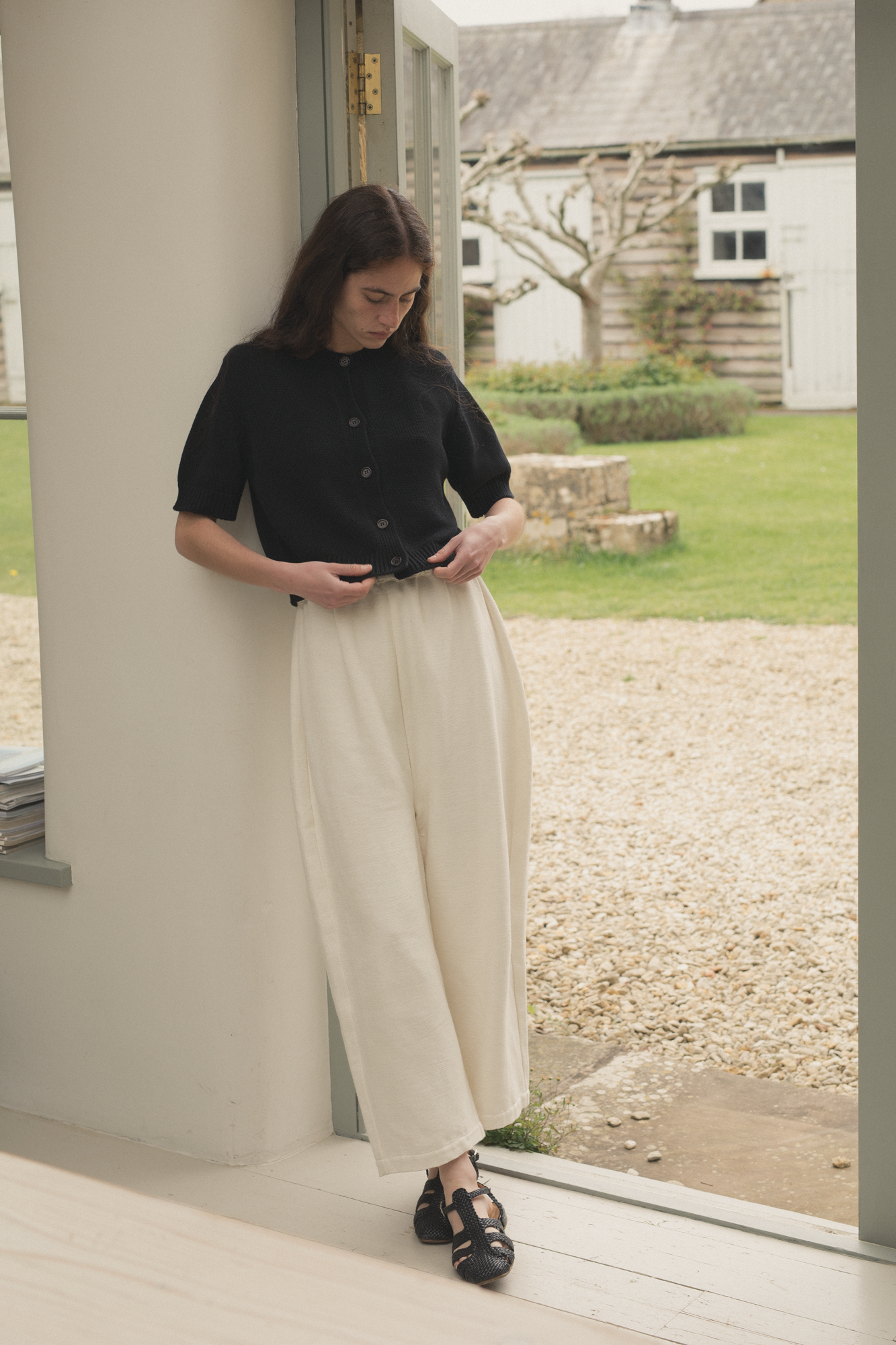Woman standing on a porch looking down with a garden and house in the background wearing the rosemary cardigan in noir by gillian stevens