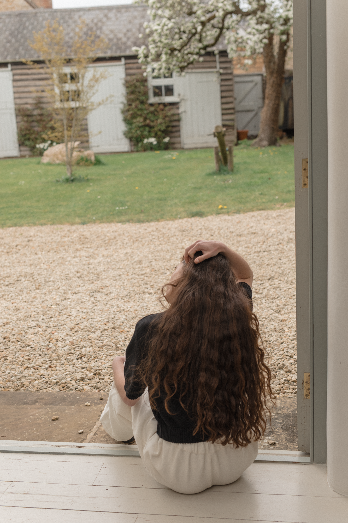 Woman sitting on a gravel path looking out of a window at a garden wearing the rosemary cardigan in noir by Gillian stevens