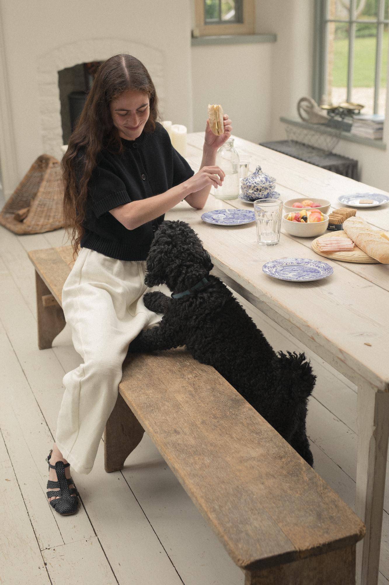 Model wearing the Rosemary Cardigan in Noir by Gillian Stevens, seated at a rustic dining table with a black dog