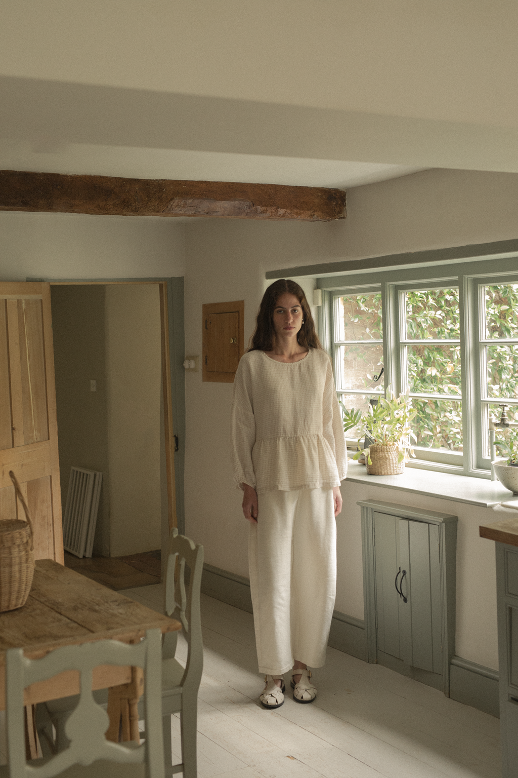 Model wearing the Darla Blouse in Oat Plaid by Gillian Stevens, full-length front view in a rustic kitchen setting