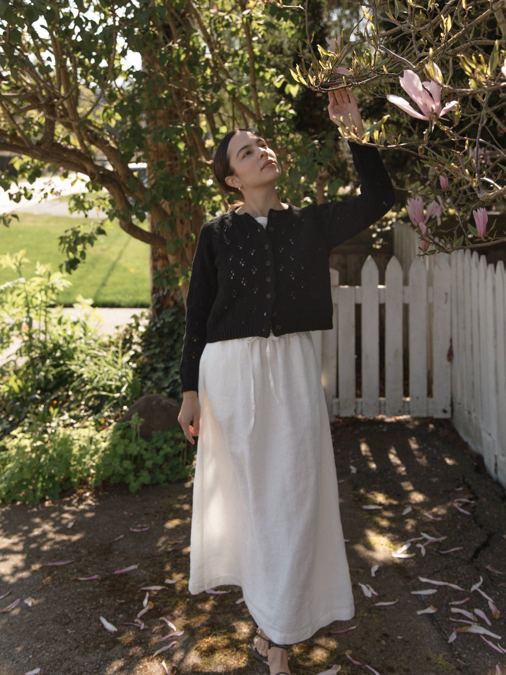 Model wearing the Mabel Cardigan in Noir by Gillian Stevens, reaching up to magnolia blooms in a garden