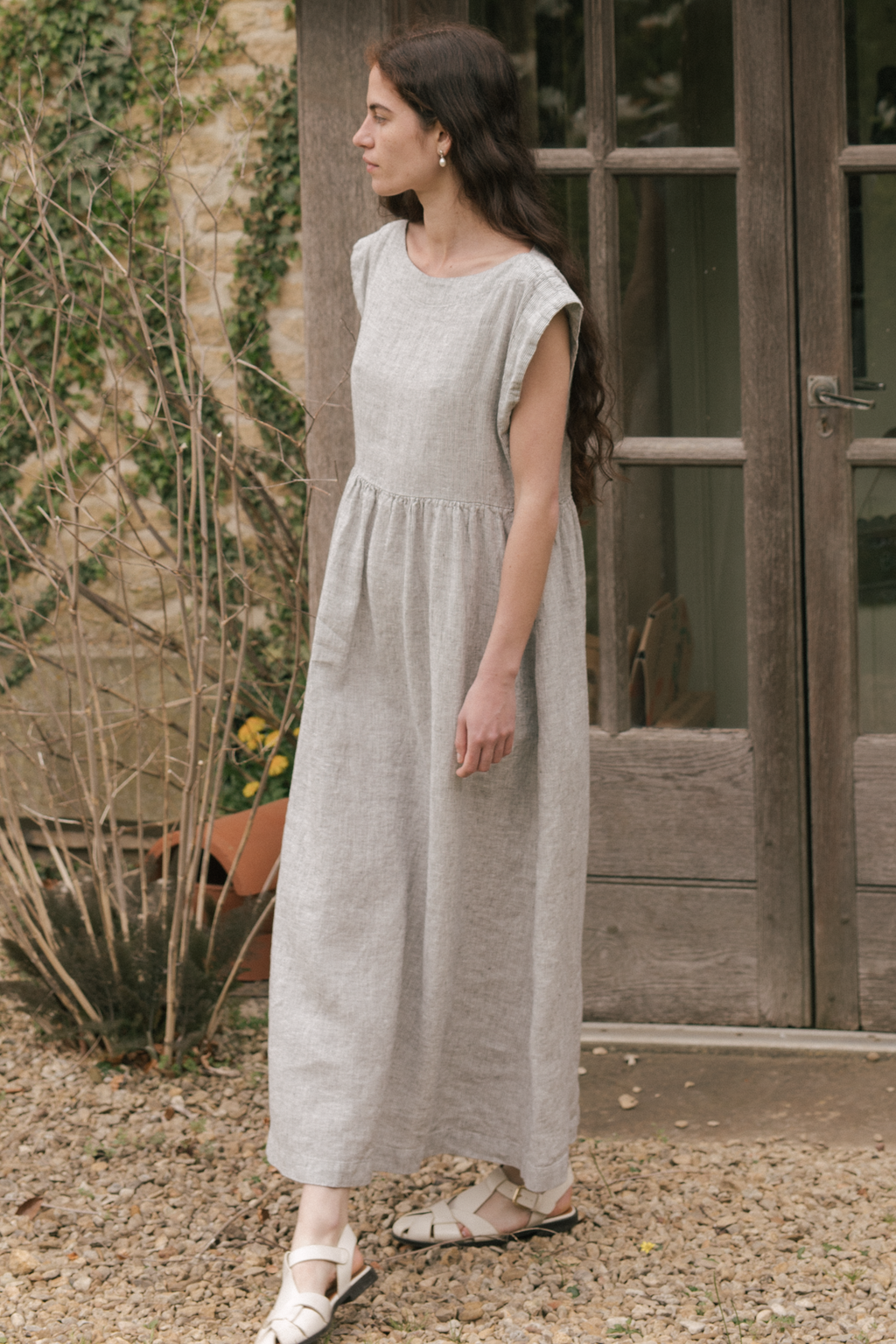 Woman in a light gray dress, the Valley Dress in Charcoal Stripe by Gillian Stevens, standing outdoors near a wooden door.
