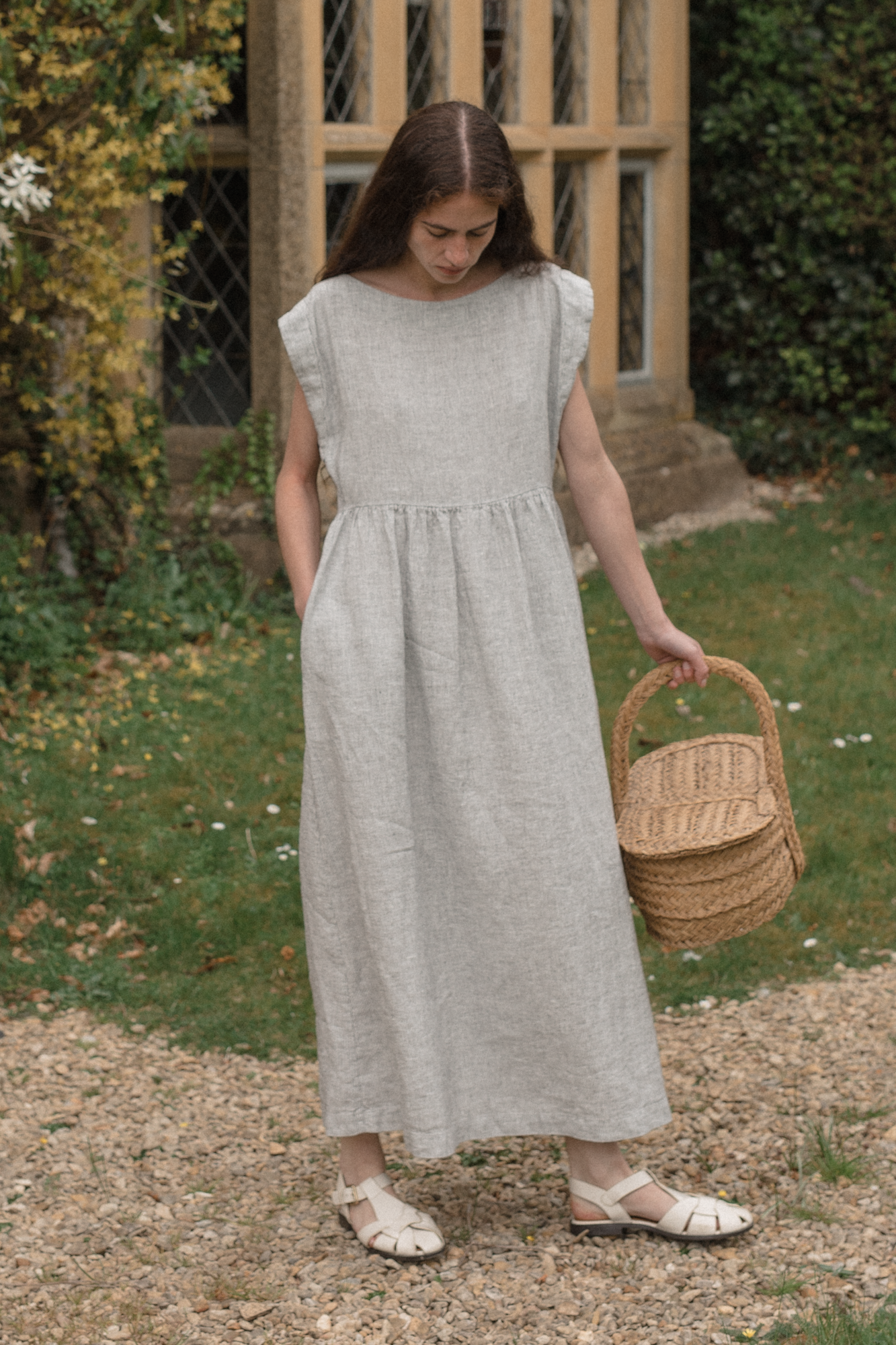 Woman in a long light gray dress, the Valley dress in Charcoal Stripe by Gillian stevens, holding a woven basket in an outdoor setting.