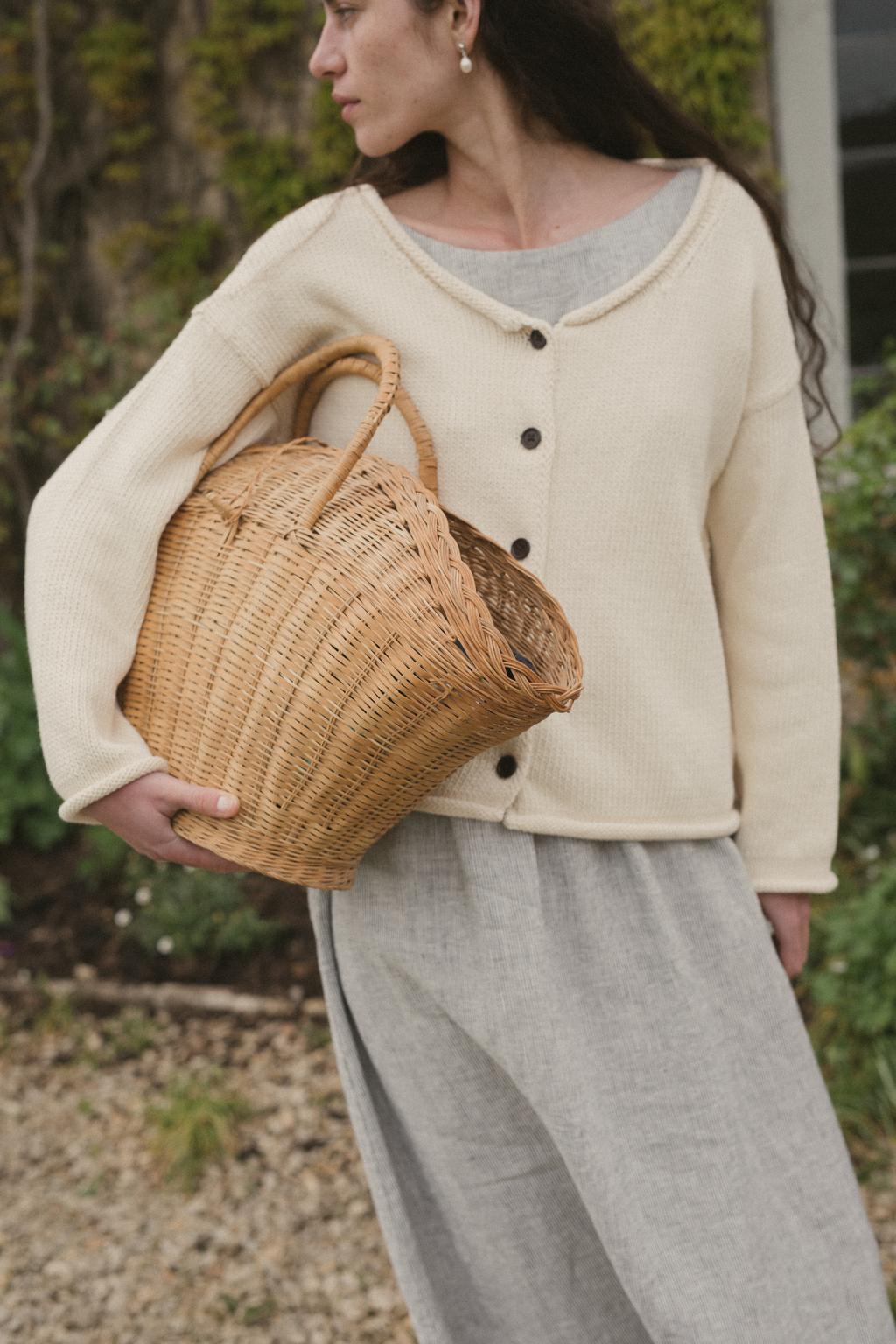 Woman holding a woven basket outdoors wearing an off white knit sweater, the Lennon Cardigan in Ivory by Gillian Stevens