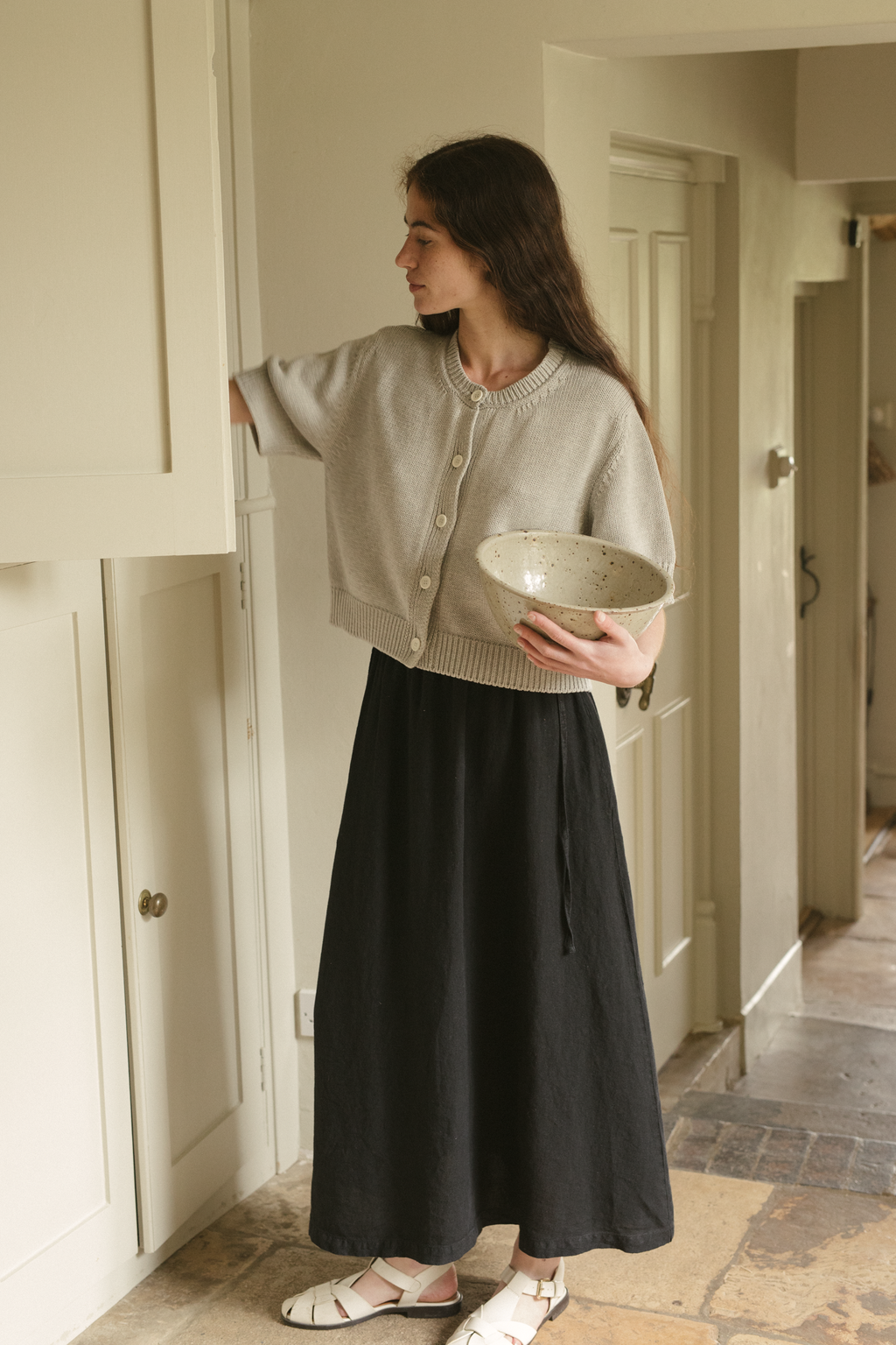 Woman holding a bowl in a home setting, wearing the rosemary cardigan in pebble by Gillian Stevens