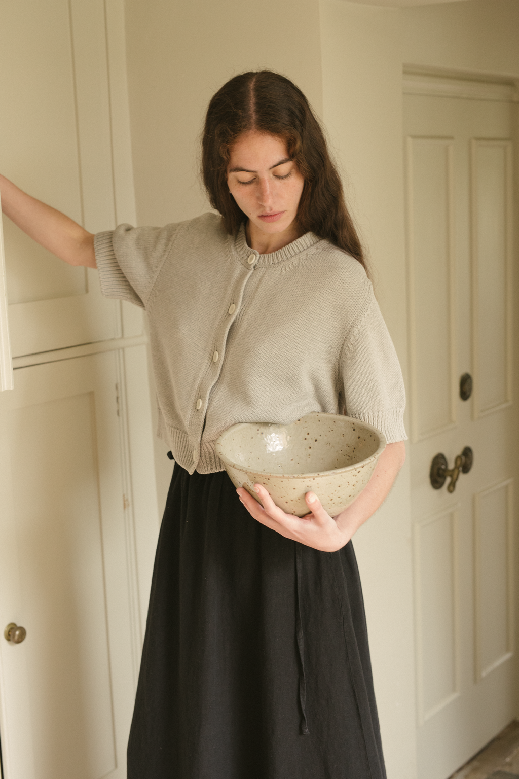 Woman holding a ceramic bowl indoors wearing the rosemary cardigan in pebble by Gillian Stevens. 