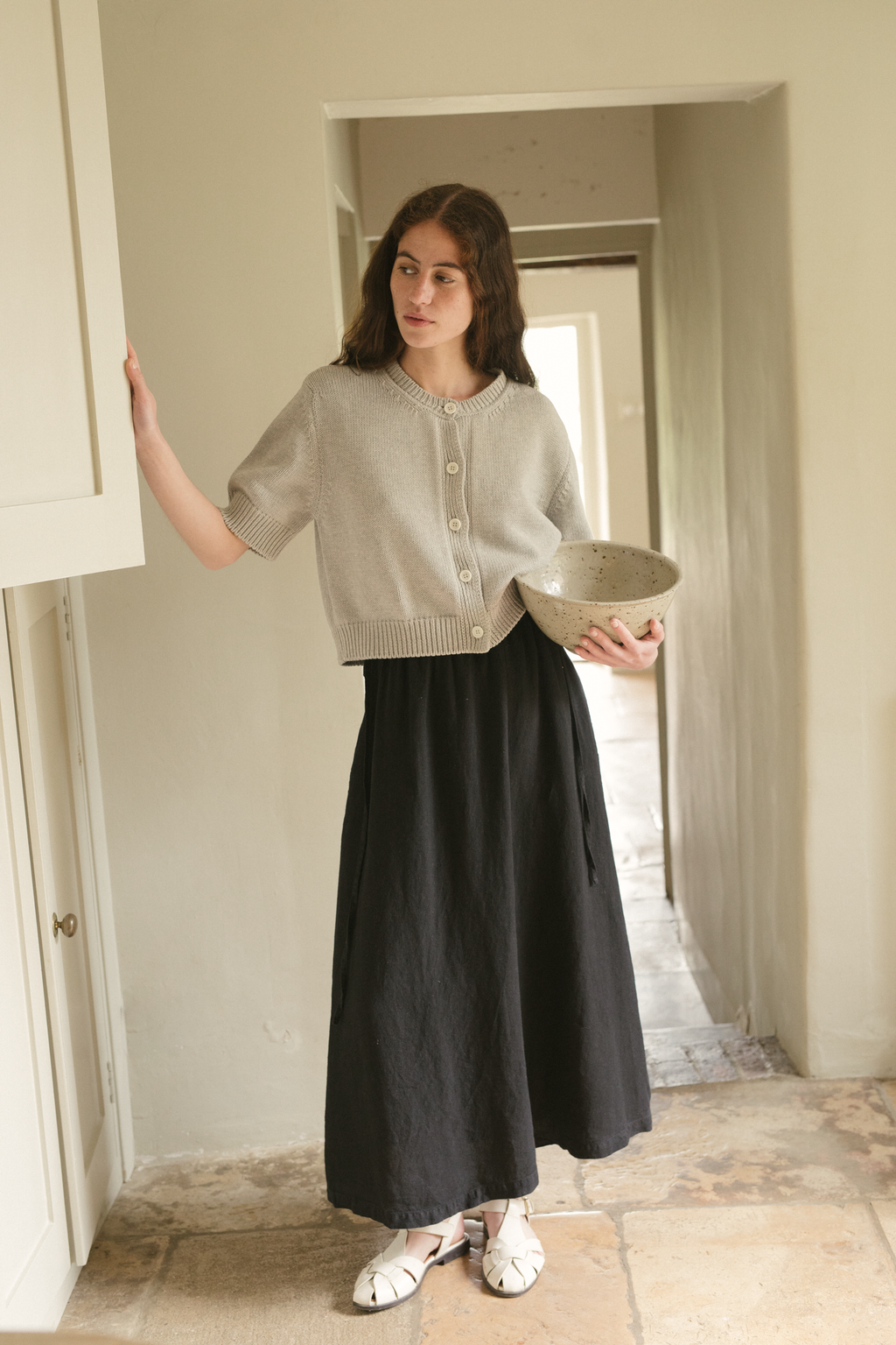 Woman holding a plate in a room with a white wall and tiled floor, wearing the rosemary cardigan in pebble by Gillian Stevens