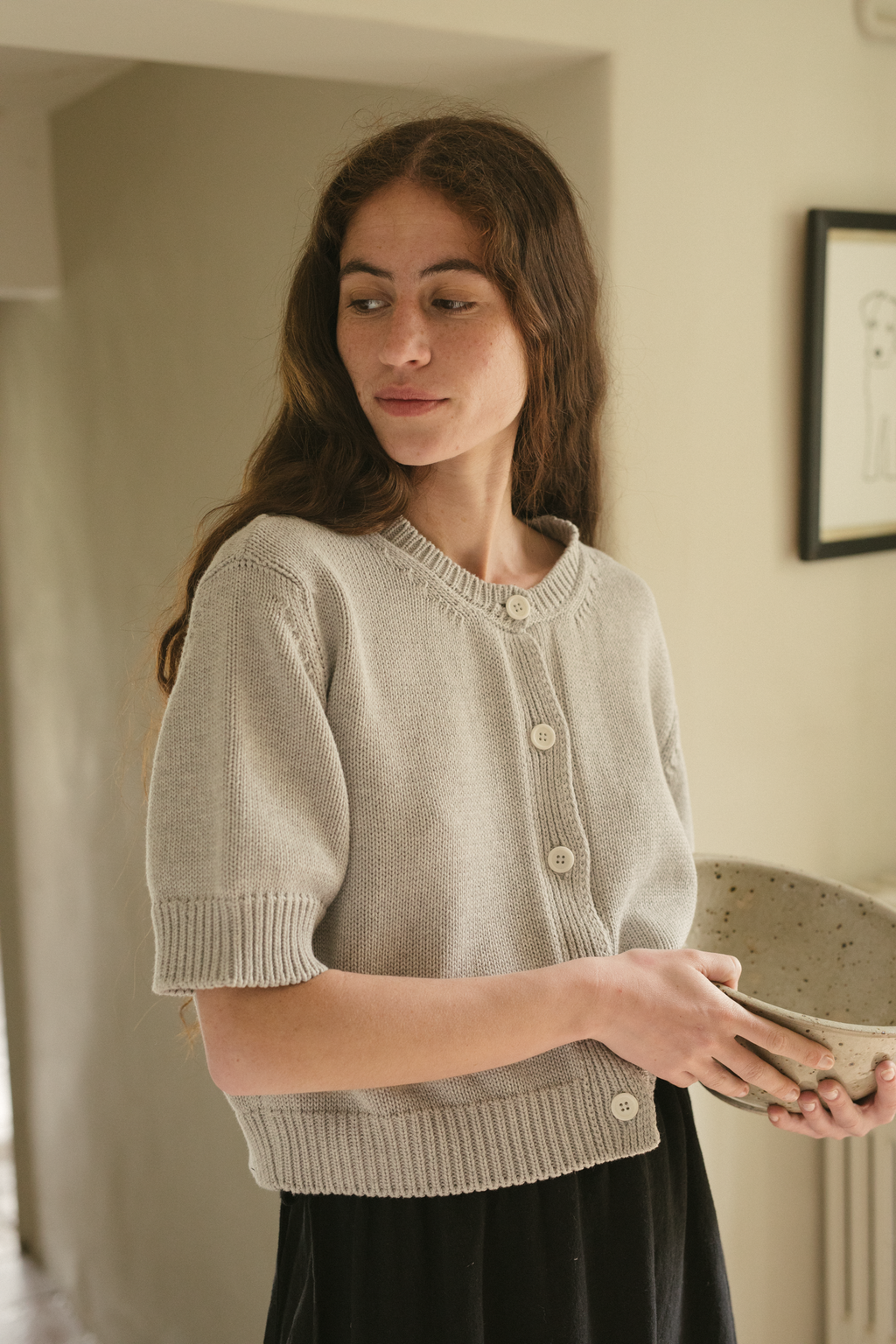 Woman wearing a light gray knit cardigan, the rosemary cardigan in pebble by Gillian Stevens, holding a ceramic bowl indoors.