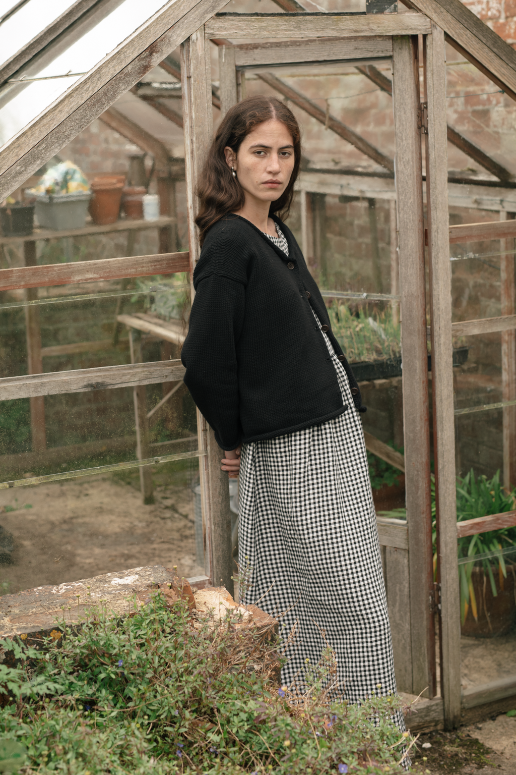 Model wearing the Lennon Cardigan in Noir by Gillian Stevens, side view in a greenhouse setting