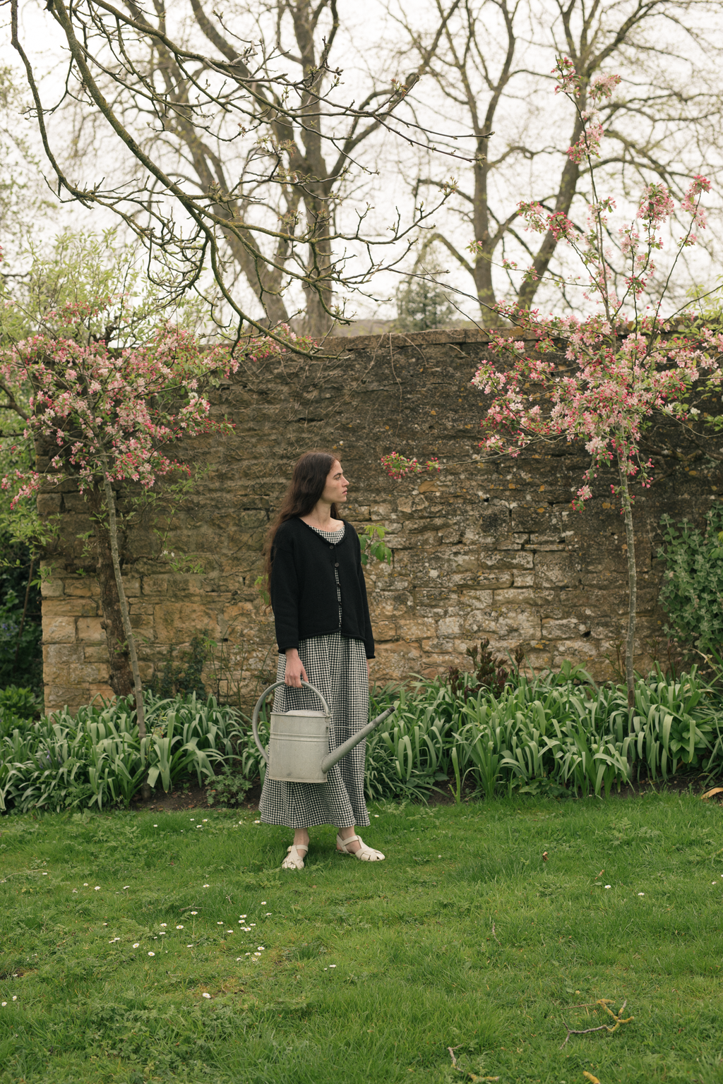 Model wearing the Lennon Cardigan in Noir by Gillian Stevens, full-length side view in a walled garden