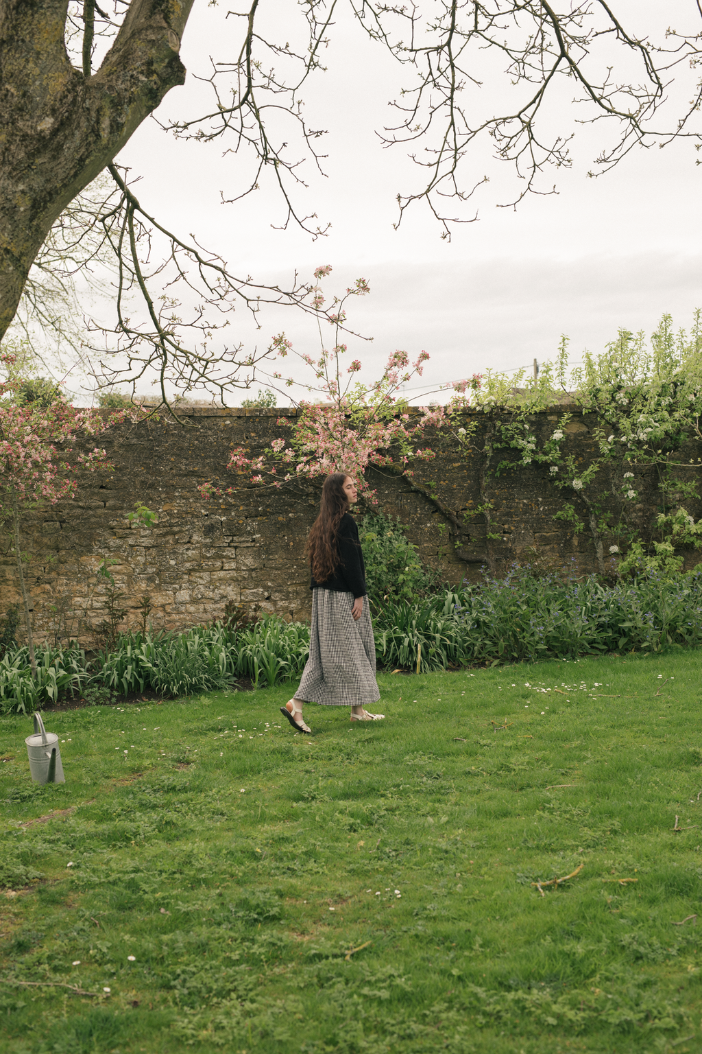 Model wearing the Lennon Cardigan in Noir by Gillian Stevens, side view walking in a walled garden