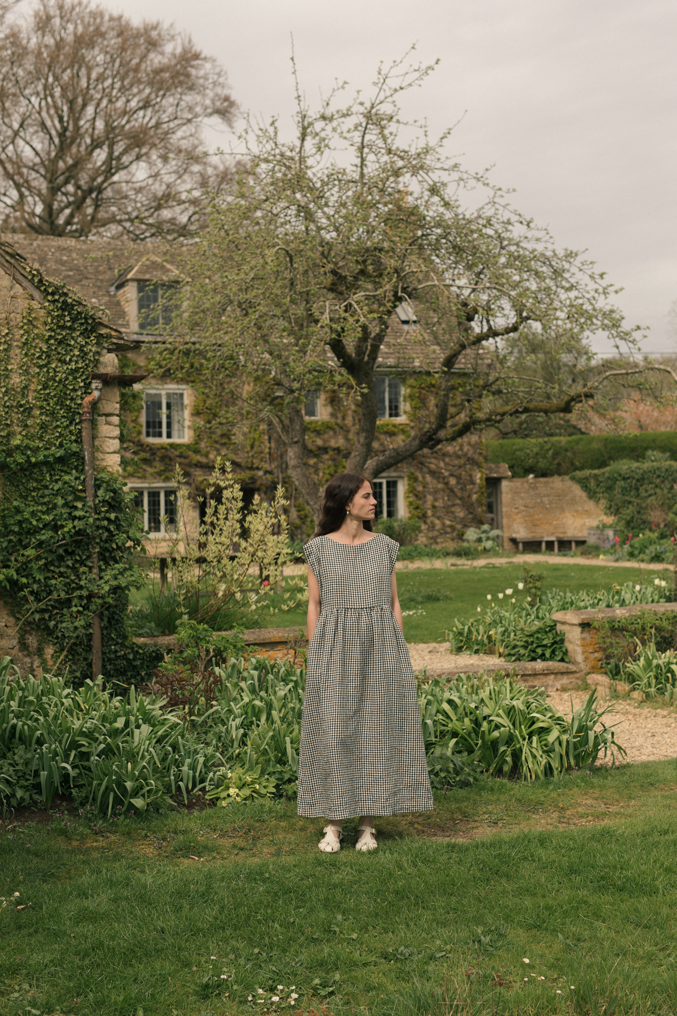 Woman in a black and white dress, the Valley Dress in Gingham noir by Gillian Stevens, standing in a garden with a stone building in the background