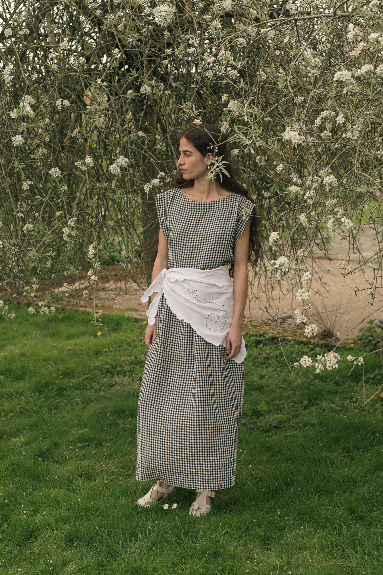 Woman in a checkered dress, the valley dress in gingham noir by Gillian Stevens, standing in a garden with white flowers and green grass.
