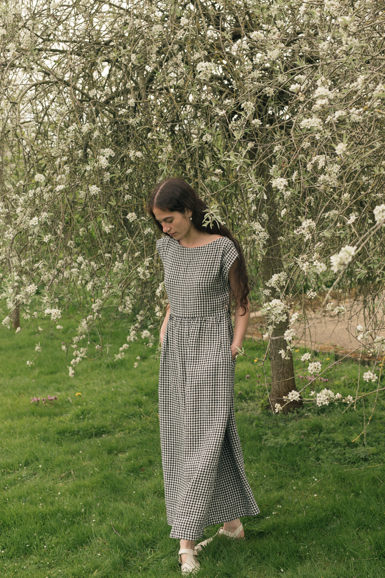 Woman in a checkered dress, the valley dress in gingham noir by Gillian Stevens,  standing in a garden with blooming trees.