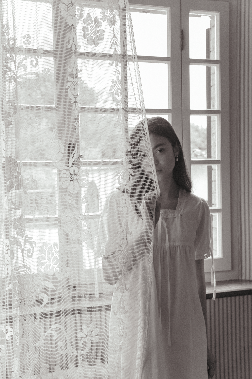 Model in the Dace Dress in Salt by Gillian Stevens, partially obscured by a lace curtain beside a window