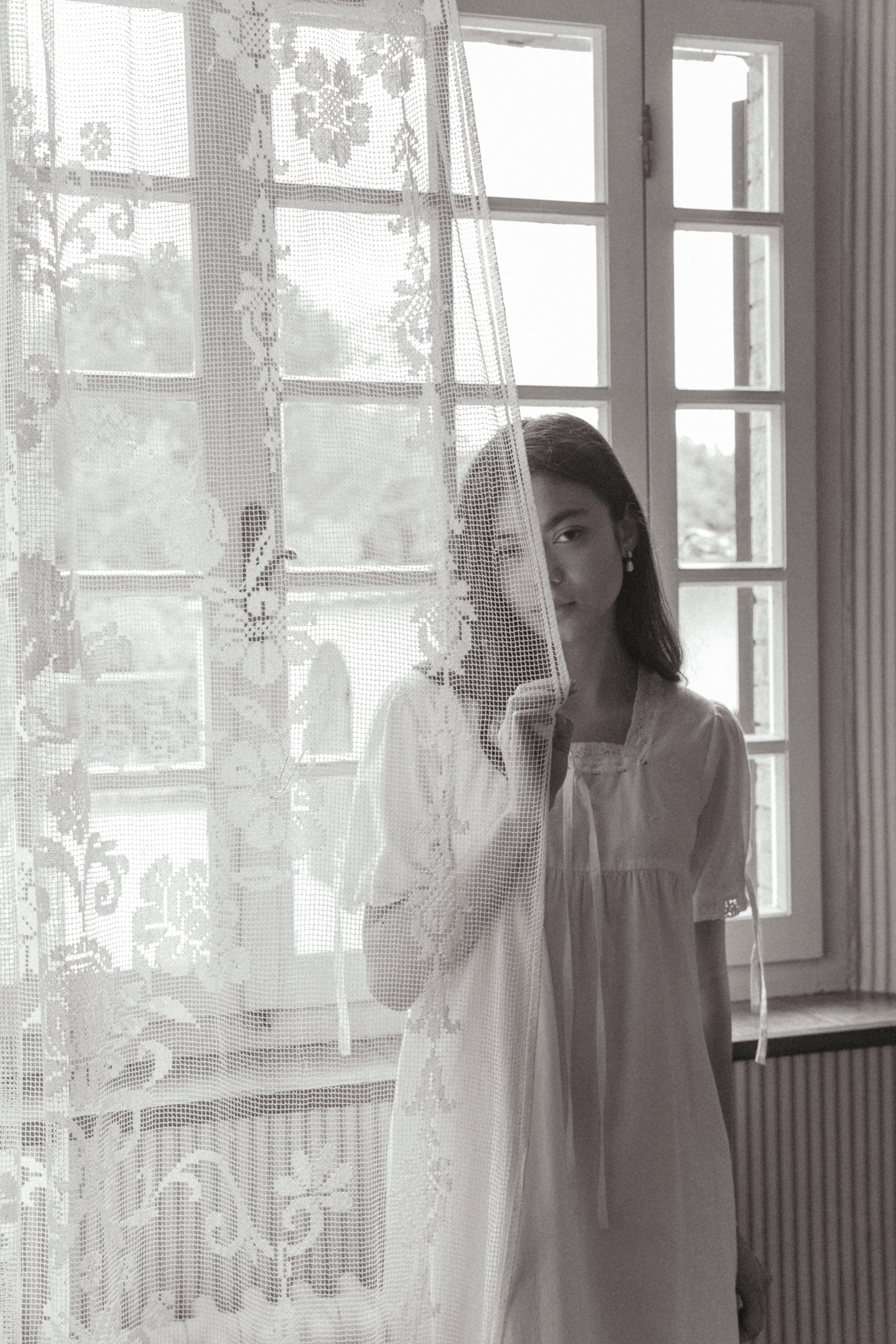 Model in the Dace Dress in Salt by Gillian Stevens, partially obscured by a lace curtain beside a window