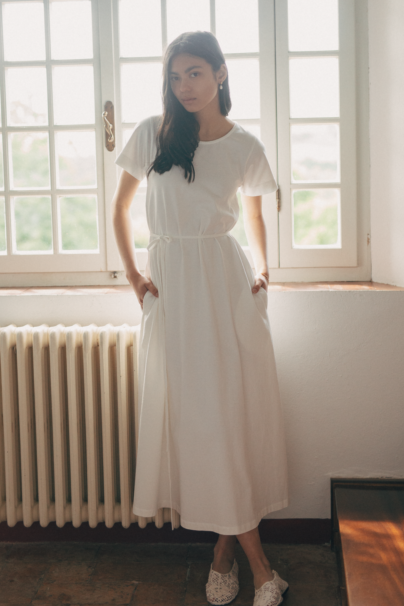 Model wearing the Palmer Dress in Salt by Gillian Stevens, front view beside a sunlit window with hands in pockets