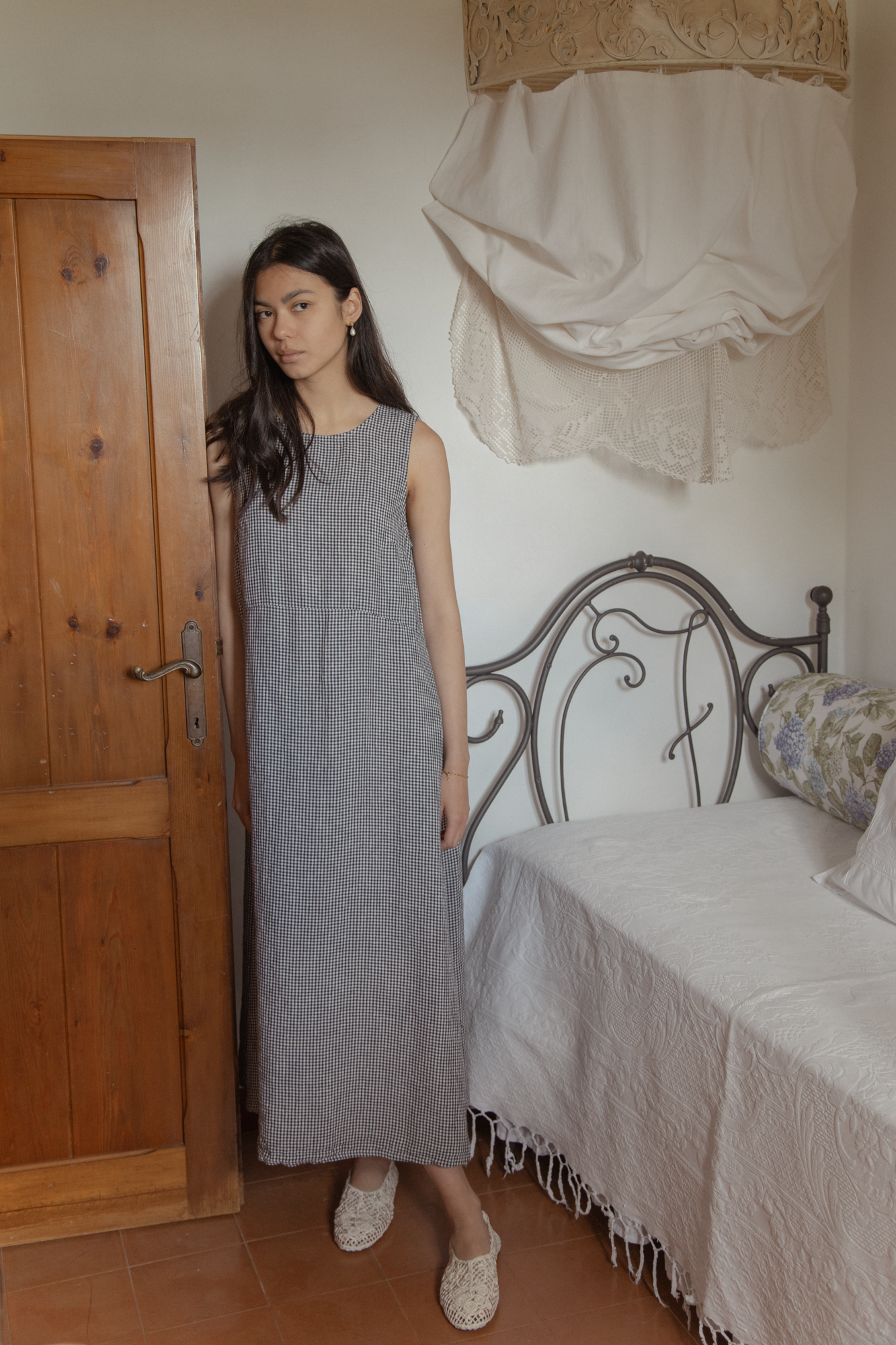 Model wearing the Paige Dress in Marine Gingham by Gillian Stevens, front view in a rustic bedroom setting