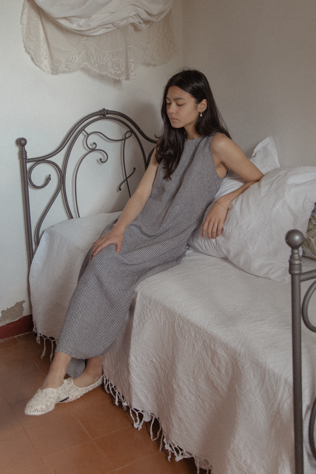 Model seated on an iron bed wearing the Paige Dress in Marine Gingham by Gillian Stevens