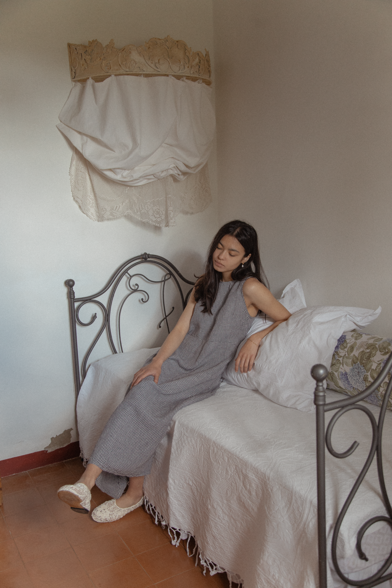 Model seated on an iron bed wearing the Paige Dress in Marine Gingham by Gillian Stevens, full-length view