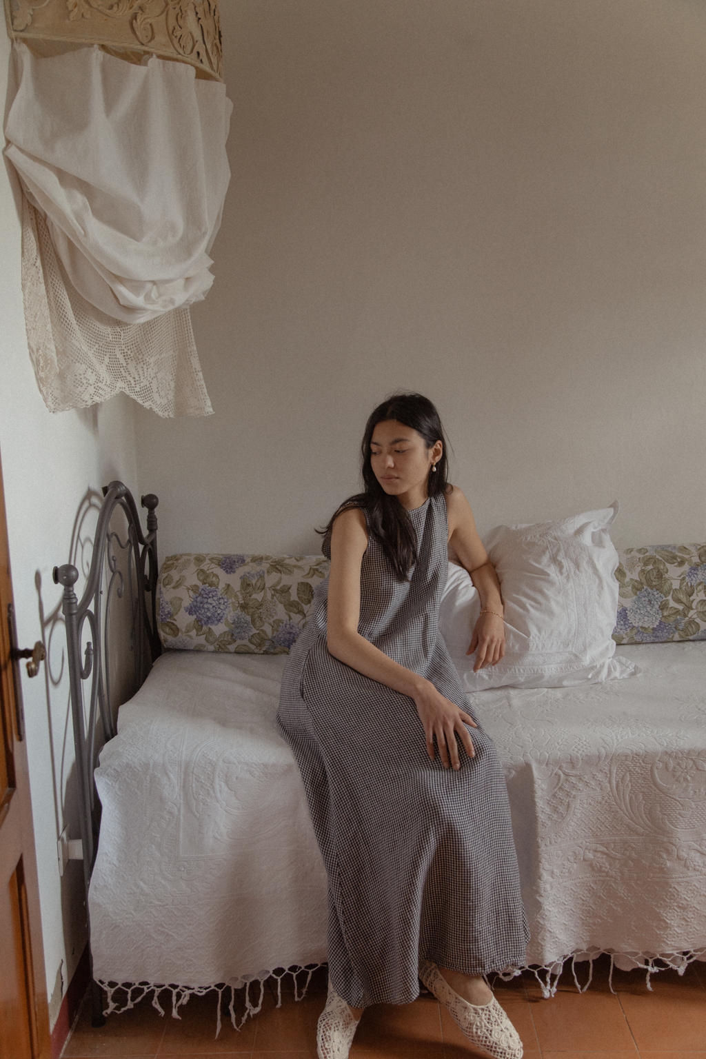 Model seated on an iron bed wearing the Paige Dress in Marine Gingham by Gillian Stevens
