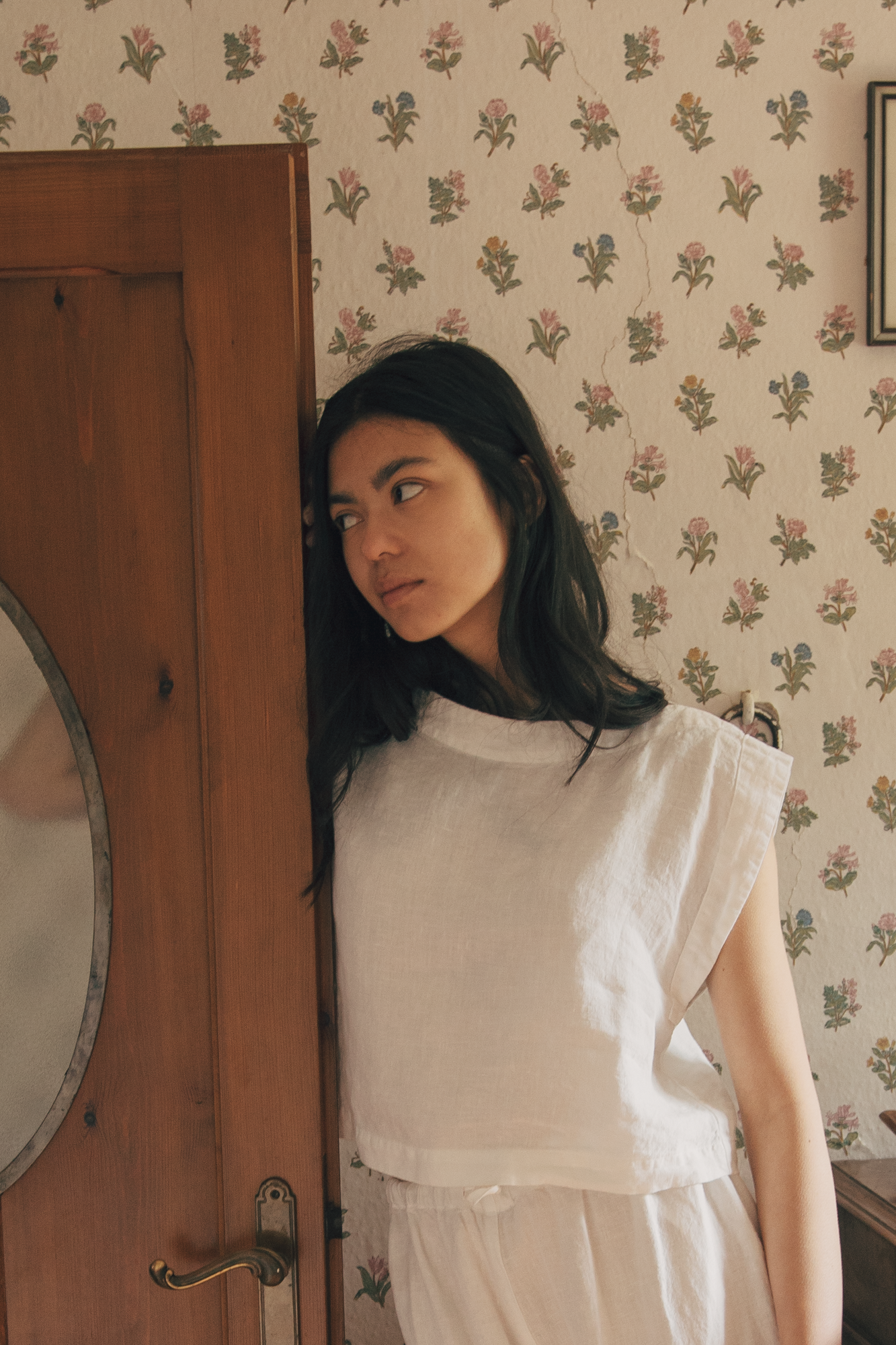 Model wearing the Valley Top in Salt by Gillian Stevens, leaning against a wooden door, floral wallpaper setting